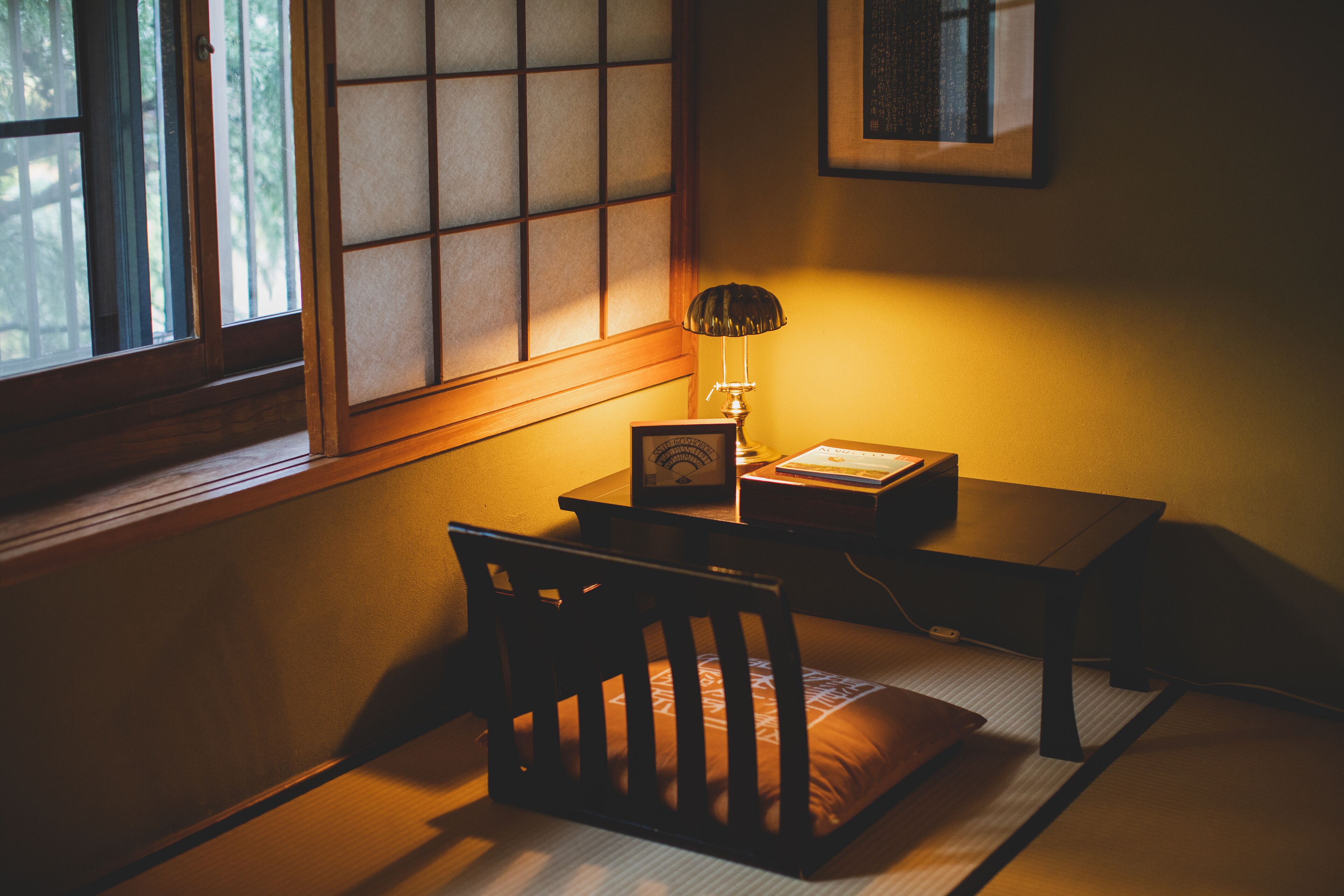 A traditional Japanese room with tatami flooring, a low wooden desk, a chair, a lamp, and a window with shoji screens.