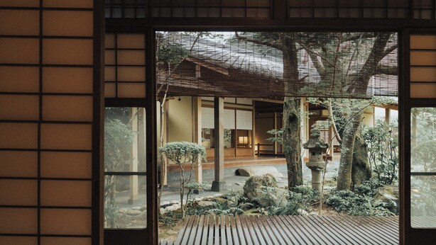 A traditional Japanese interior with tatami flooring, a shoji screen door, and a view of a landscaped garden with rocks and a wooden structure. - Tocen Goshoboh (Kobe)