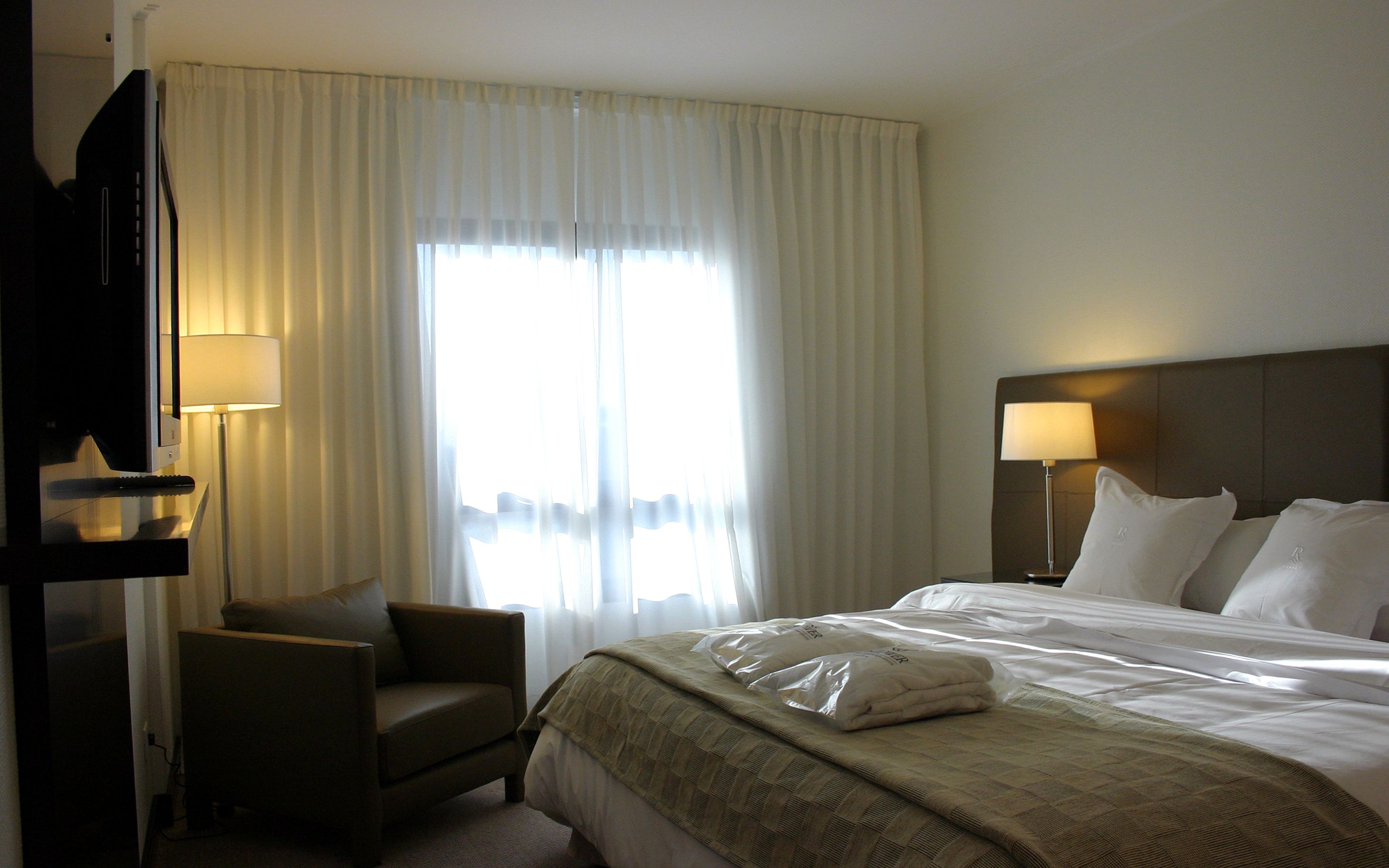 A hotel room with a bed, a chair, a desk with a lamp, and a window with curtains.