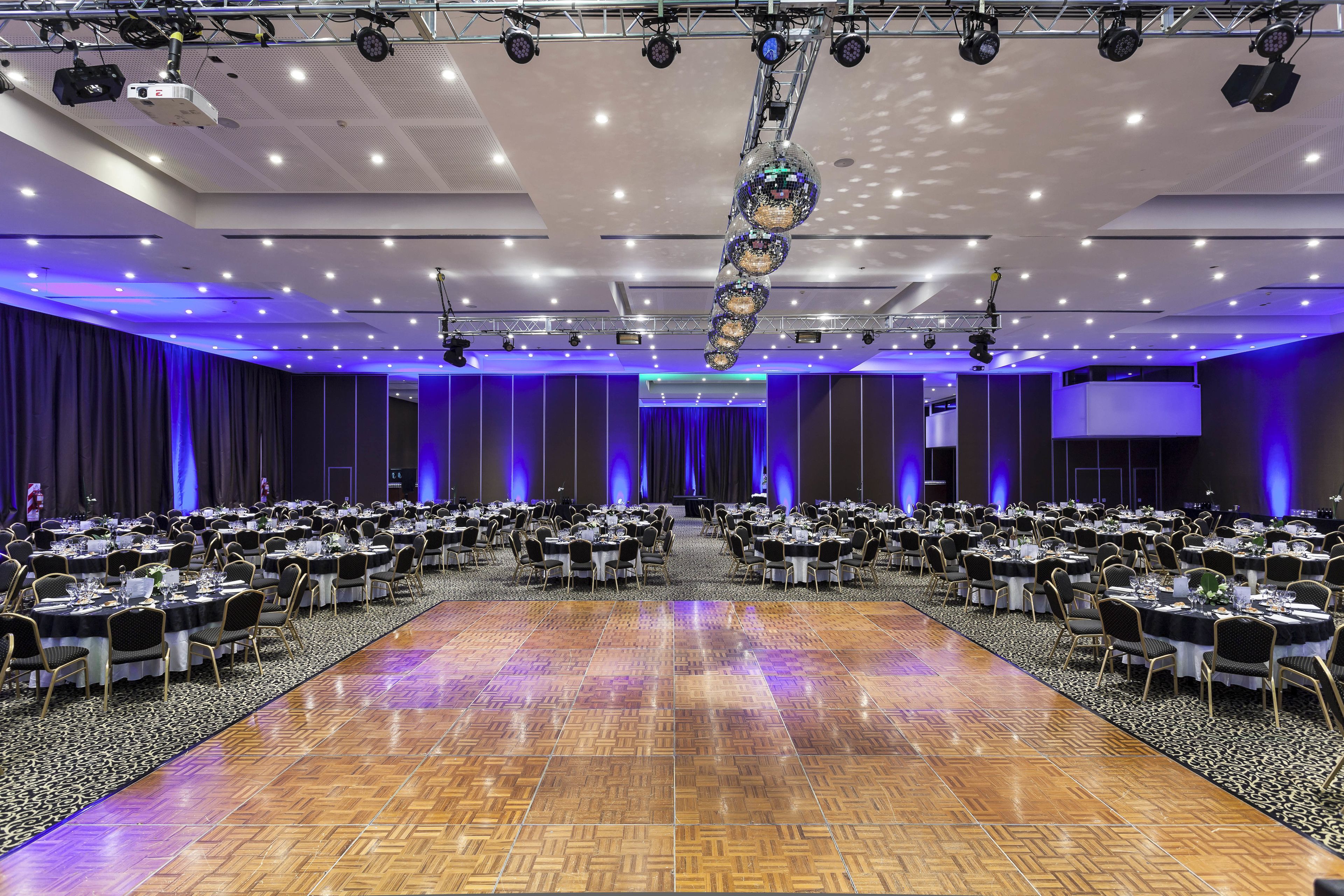 Uma sala de conferências ampla com várias mesas redondas, cadeiras e um palco com uma tela grande.