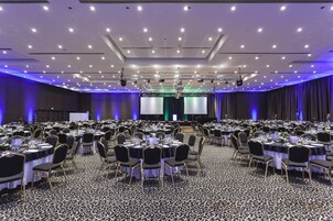 Un amplio salón de banquetes con mesas y sillas redondas, un escenario con una pantalla, y suelo alfombrado.