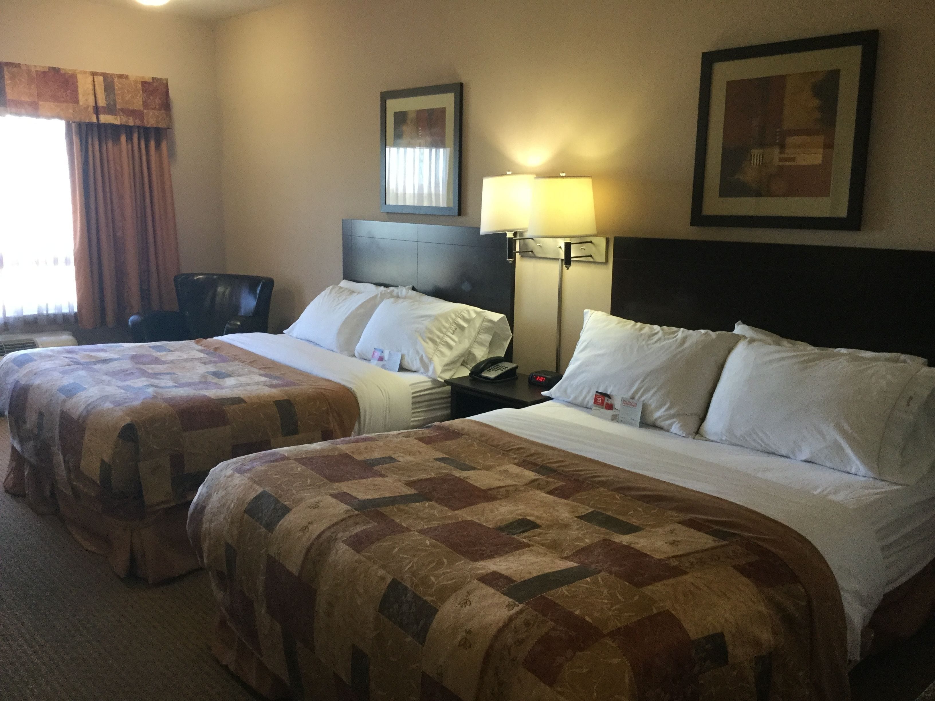 A hotel room with two beds, a nightstand with a lamp, a chair, and a framed picture on the wall.