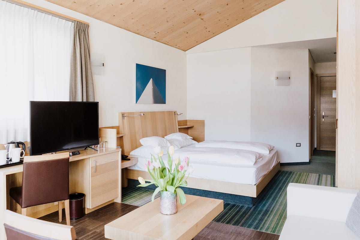 Habitación de hotel con una cama, un escritorio con televisor, una mesita con flores y un cuadro en la pared.