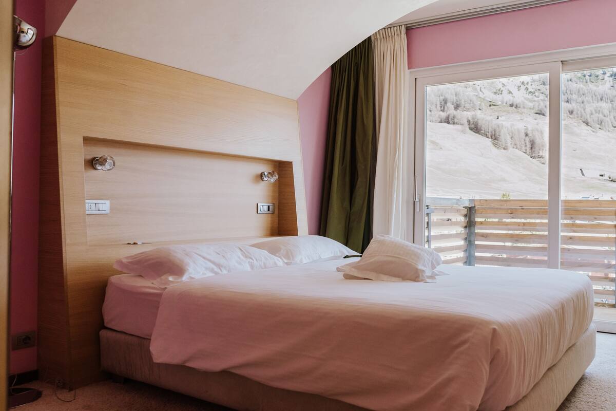 Habitación de hotel con cabecera de madera, una cama grande con ropa de cama blanca, vista a un paisaje nevado a través de una puerta corrediza de vidrio y un balcón con baranda de madera.