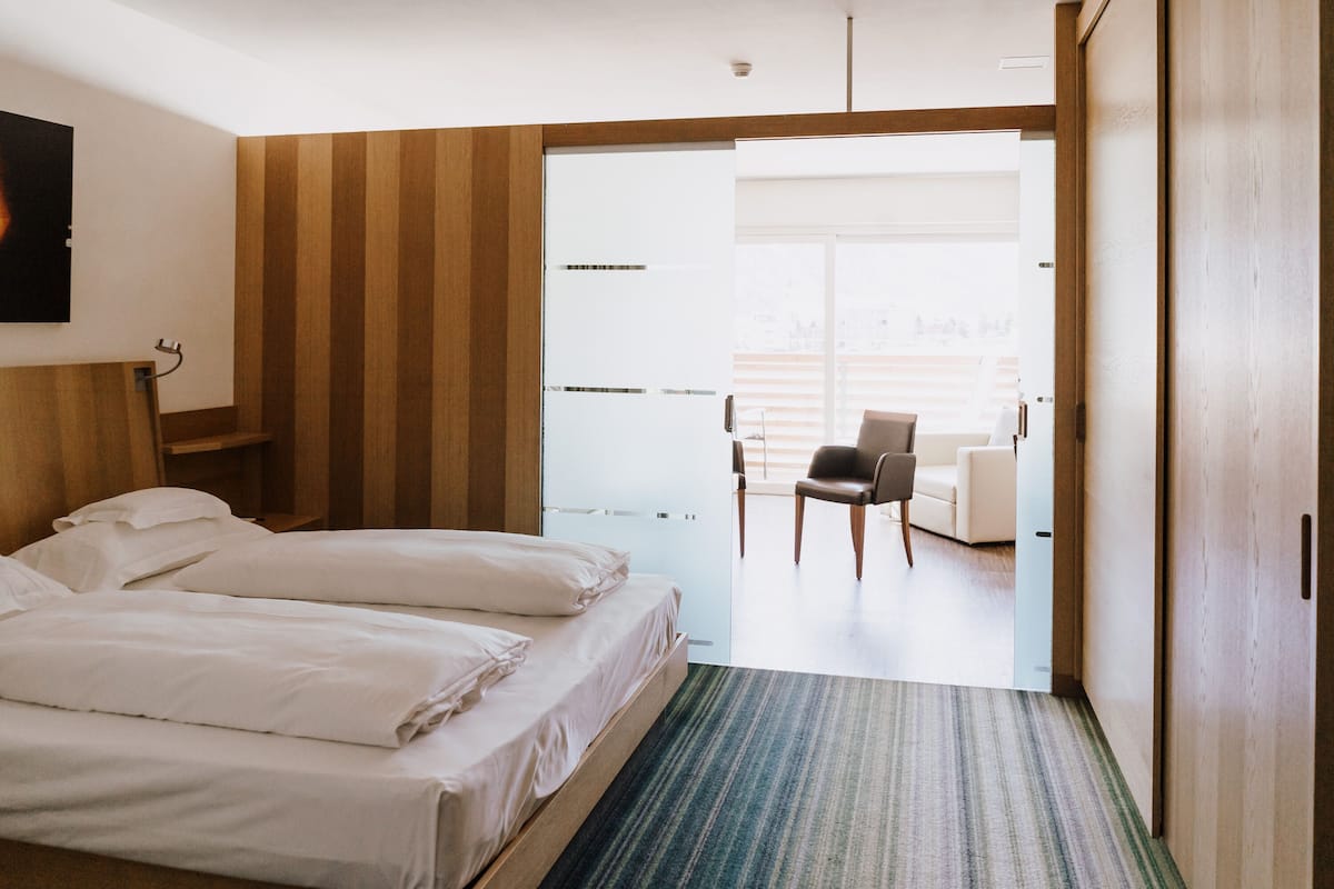 Habitación de hotel con dos camas, cabecera de madera, alfombra a rayas y vistas a un balcón con una silla.