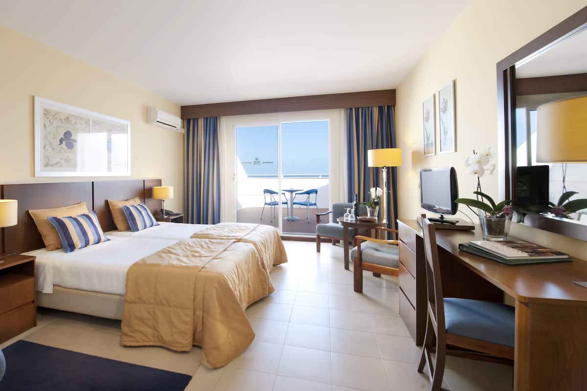 A hotel room with two beds, a desk, a chair, a TV, and a balcony with a view.