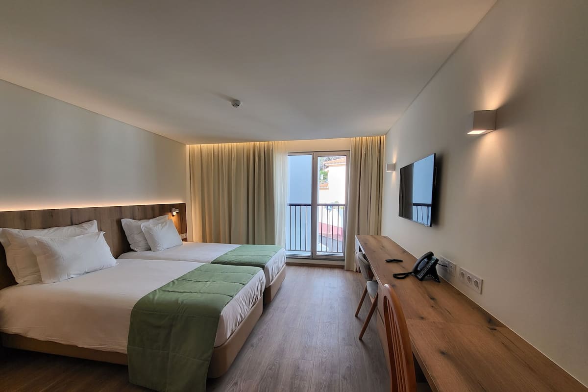 A hotel room with two beds, a desk, a chair, a TV, and a balcony with curtains.