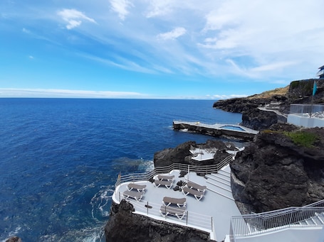A coastal cliffside with a pool area featuring sun loungers and a staircase leading down to the water.