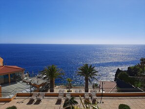 A rooftop terrace with a view of the sea, palm trees, and a building with a red roof.