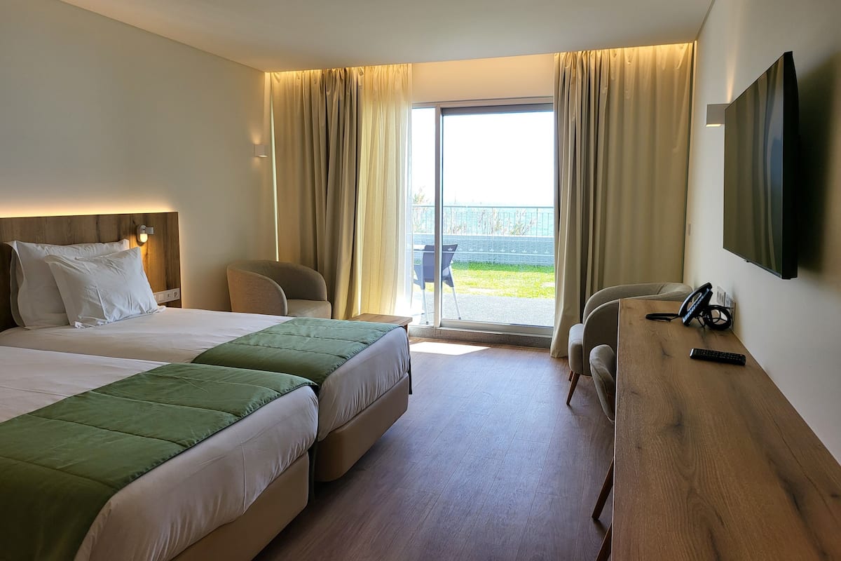 A hotel room with two beds, a desk, a chair, a TV, and a view of the outdoors.