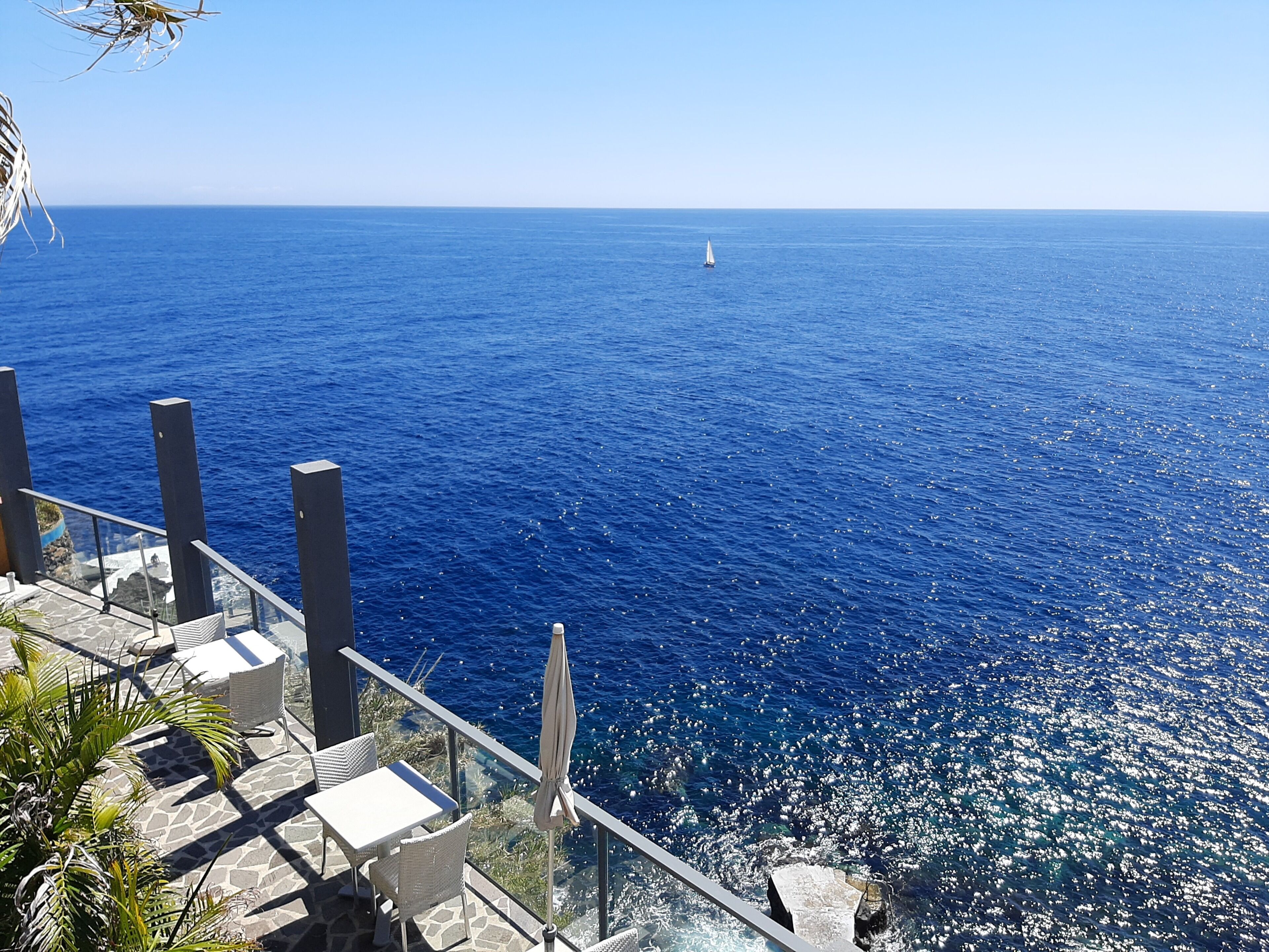 A seaside terrace with a clear view of the ocean, a sailboat in the distance, and outdoor seating.