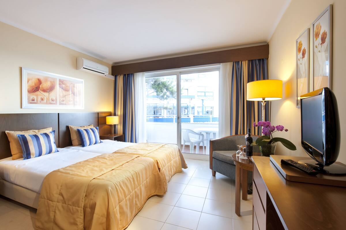 A hotel room with a bed, a desk with a TV, a chair, and a balcony with a view.