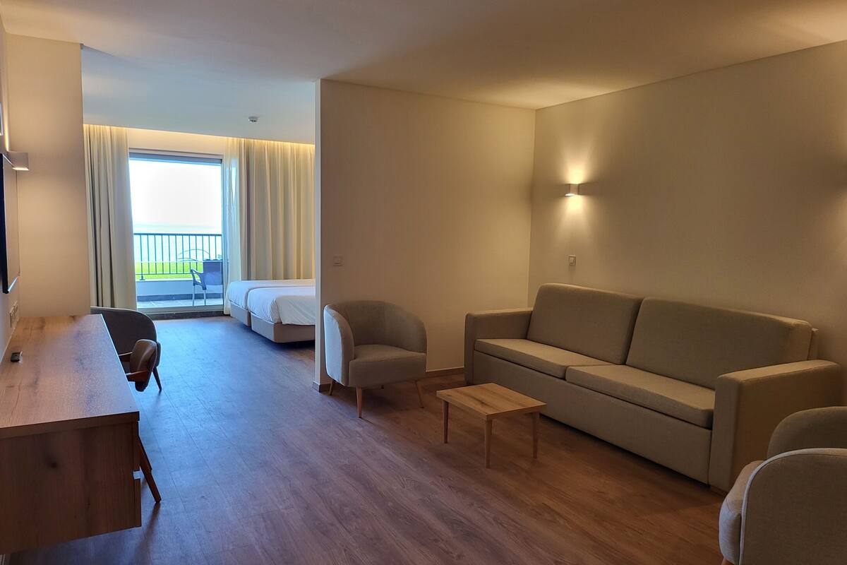 A modern hotel room with a wooden desk, two armchairs, a sofa, and a small table. A balcony with curtains is visible through the window.
