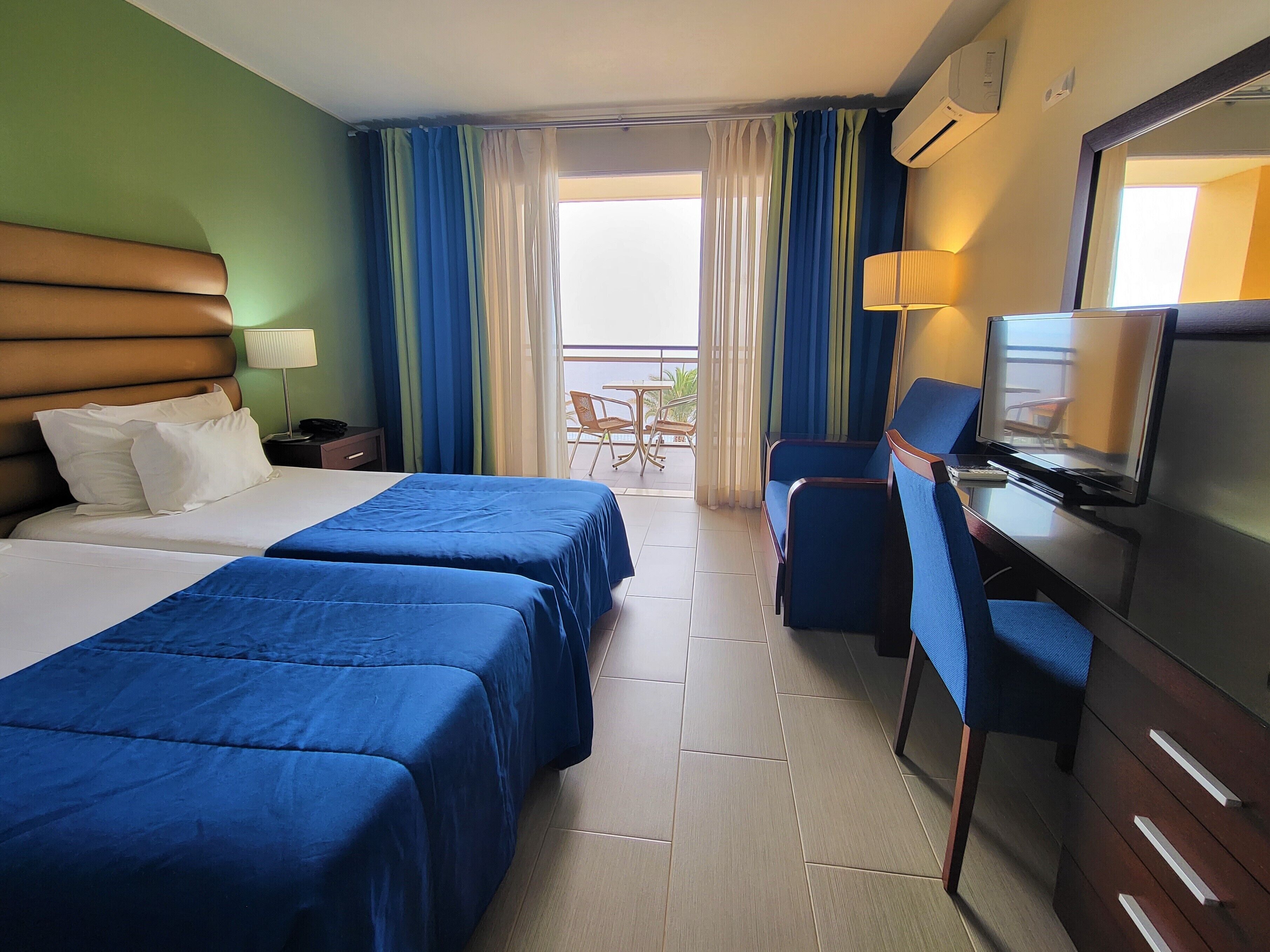 A hotel room with two beds, a desk with a television, a chair, and a balcony with a table and chairs.