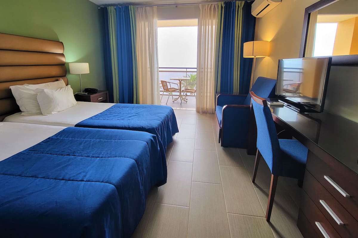 A hotel room with two beds, a desk with a television, a chair, and a balcony with a table and chairs.