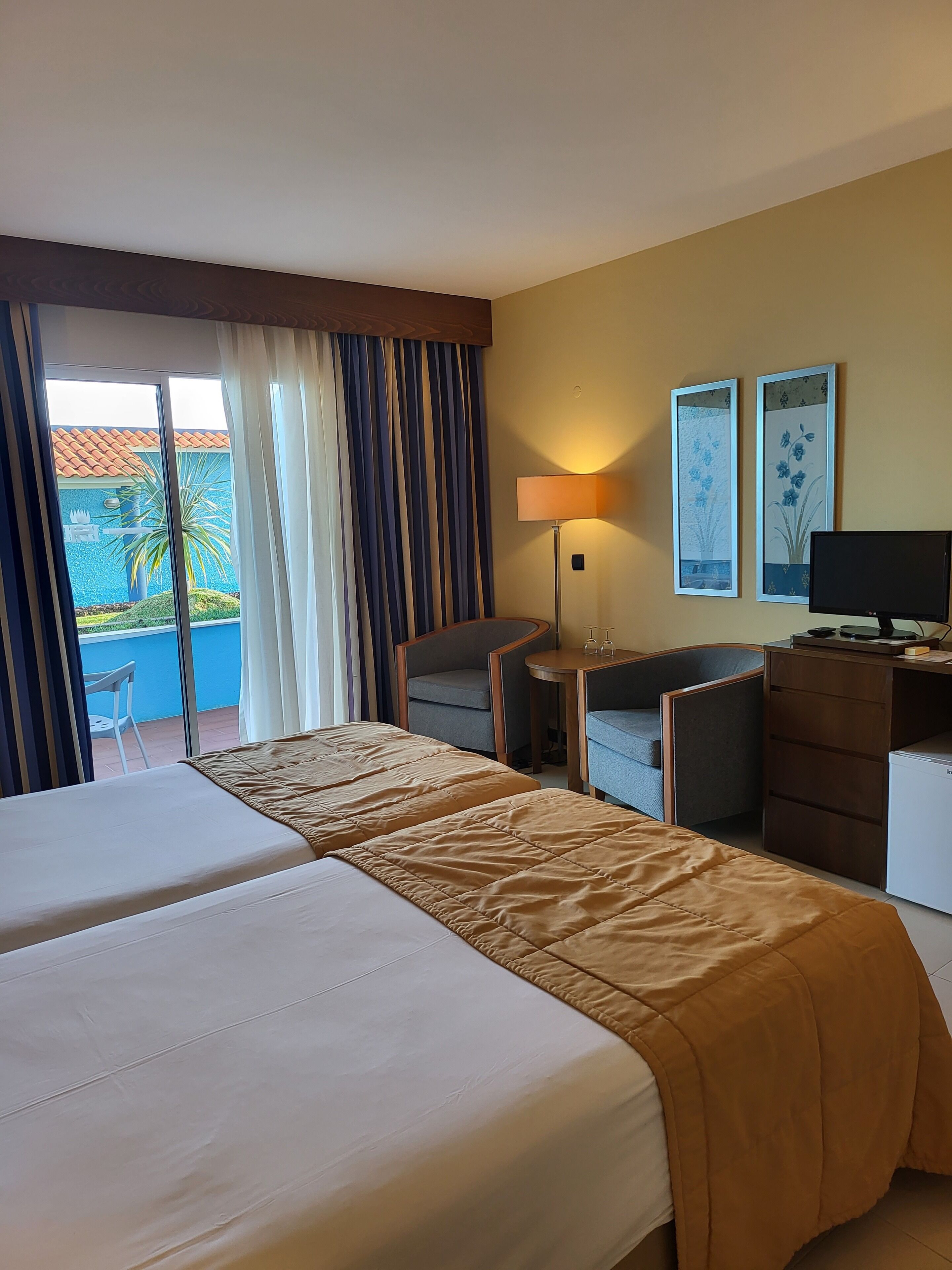 A hotel room with two beds, a desk, a chair, a lamp, and two wall-mounted paintings.