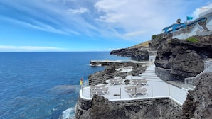 A coastal hotel with a rocky cliffside, a pool area with lounge chairs, and a view of the ocean. - Rocamar Lido Resort (Santa Cruz)