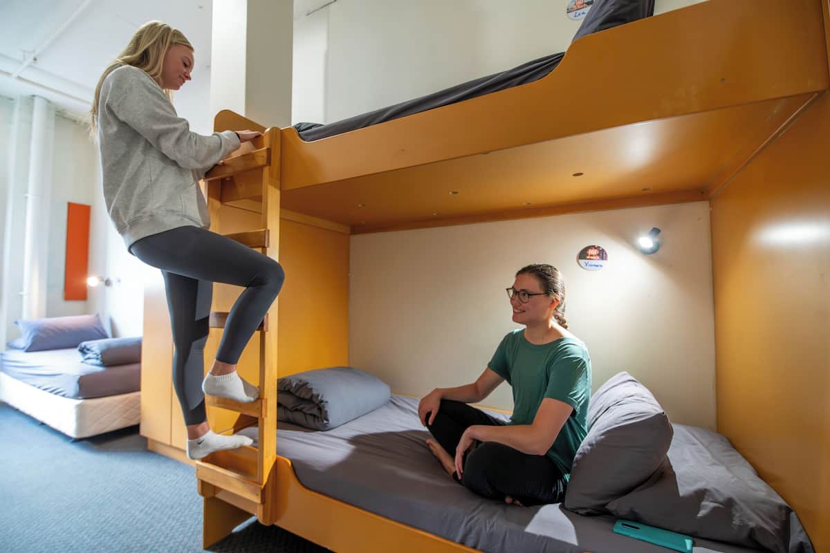 A bunk bed with two people, one standing and one sitting, in a room with a bed and a small table.