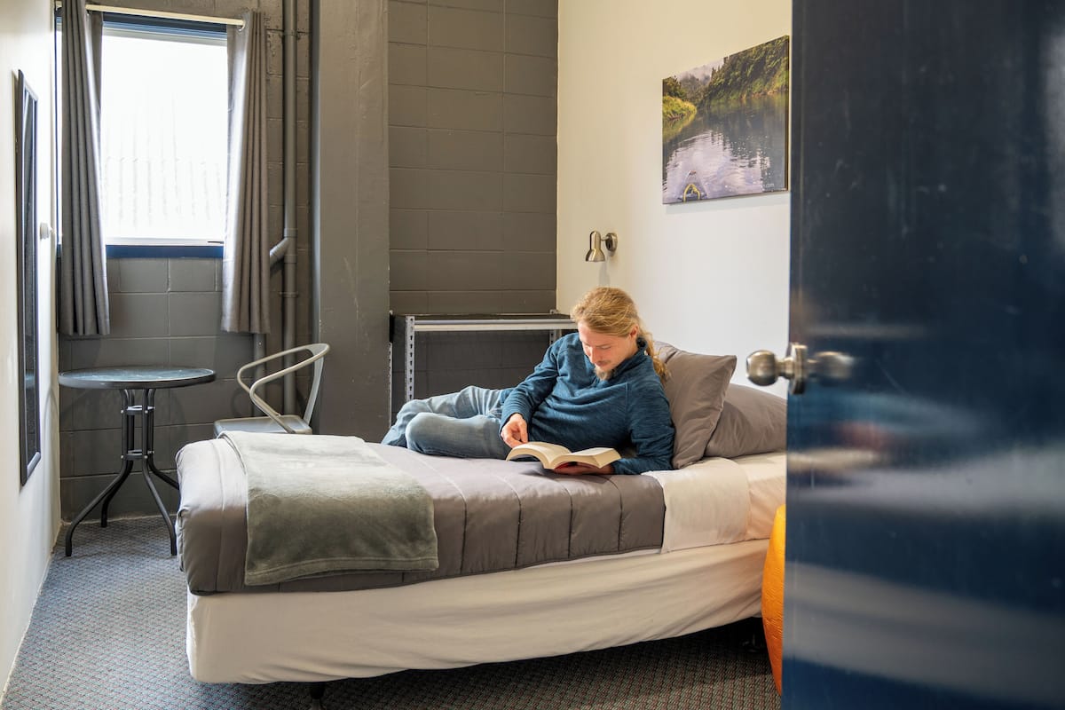 A bed with a person reading a book, a small table, a chair, and a window with curtains.