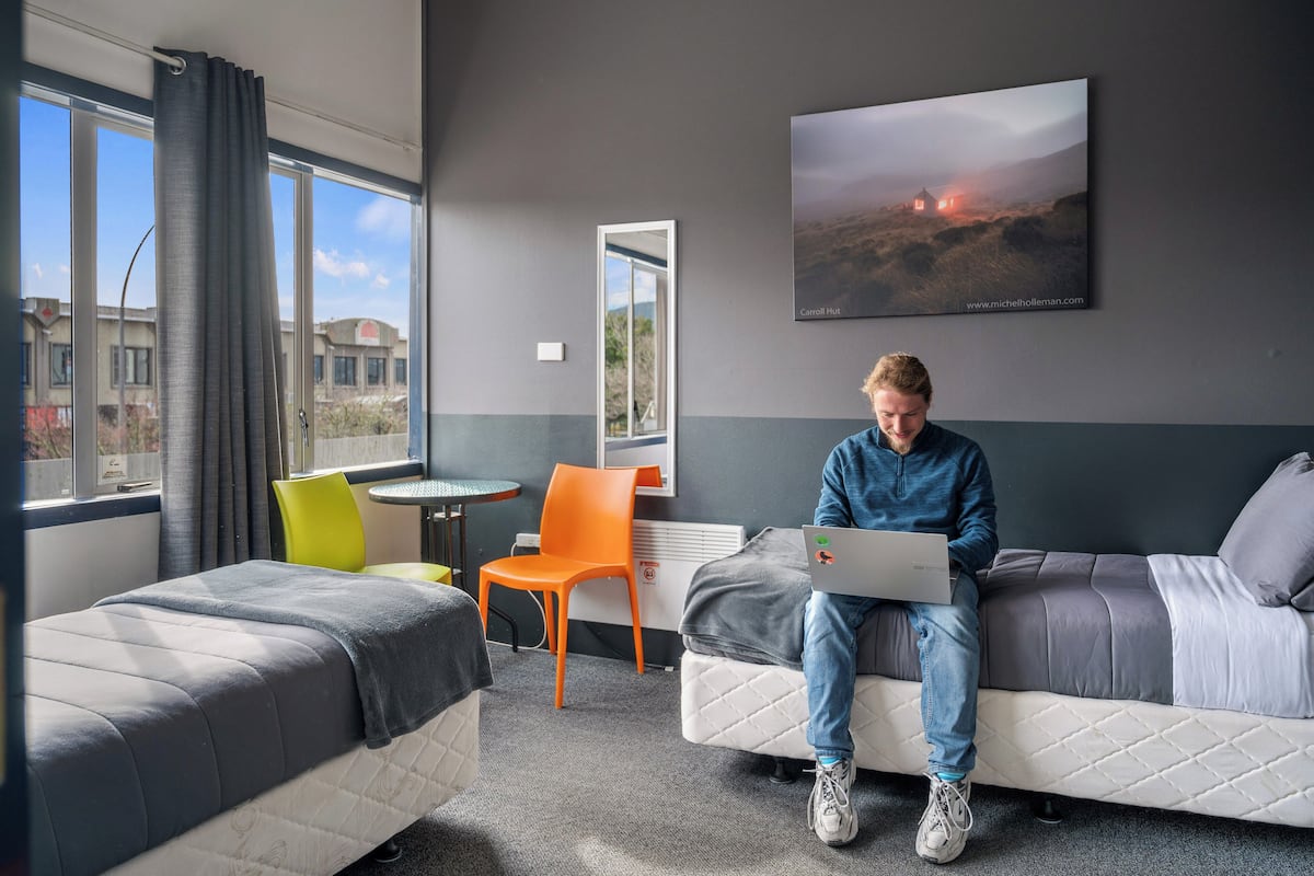 A hotel room with two beds, a desk, and a chair. There is a large window with curtains, a painting on the wall, and a view of buildings outside.