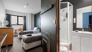 A hotel room with two beds, a desk with a chair, a small table, a fan, a shower, and a sink.