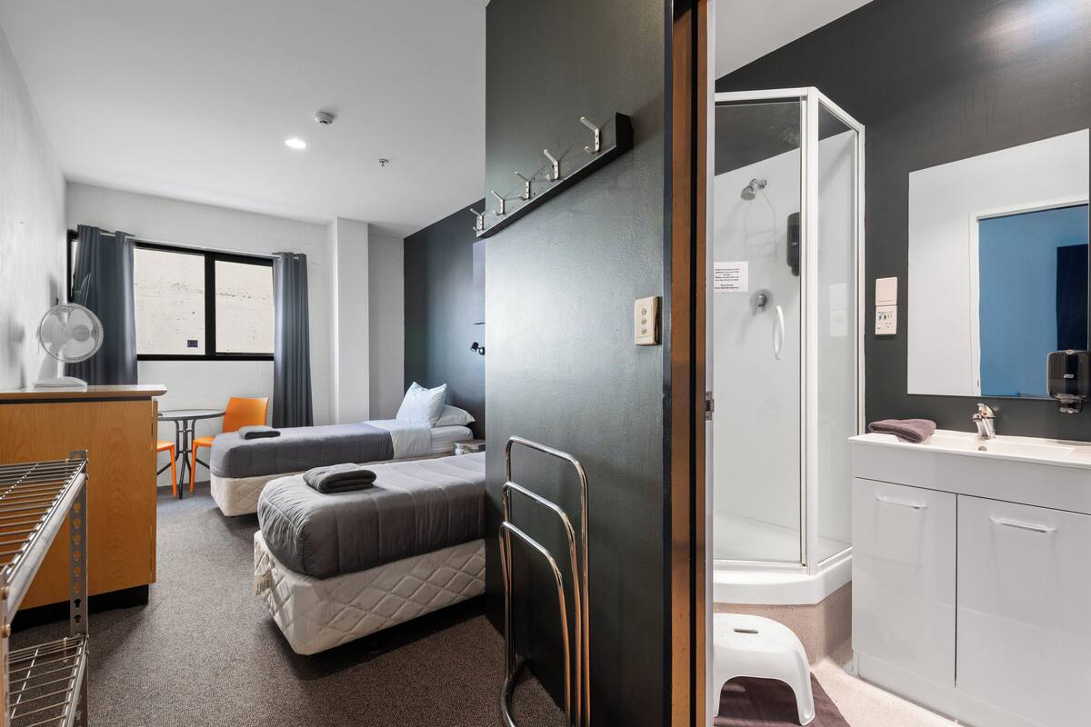 A hotel room with two beds, a desk with a chair, a small table, a fan, a shower, and a sink.