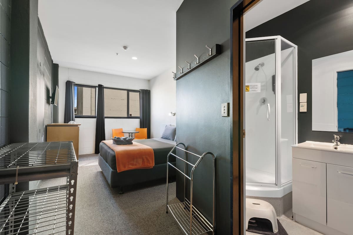 A compact hotel room with a bed, a small table, a shower area, and a kitchenette.