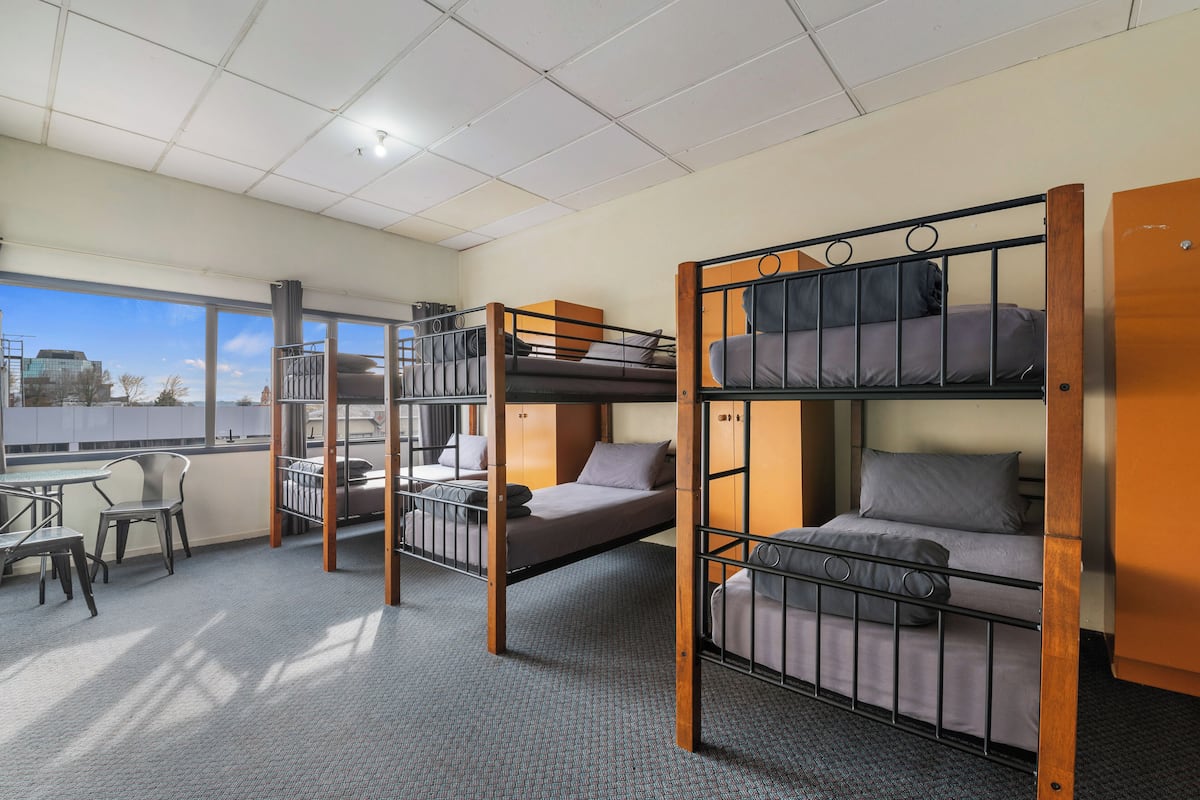 A dormitory room with bunk beds, a desk, chairs, and a large window offering a city view.