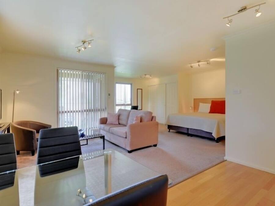 Ein modernes Hotelzimmer mit einem Glastisch, einer Couch, einem Bett und einem großen Fenster mit Jalousien.