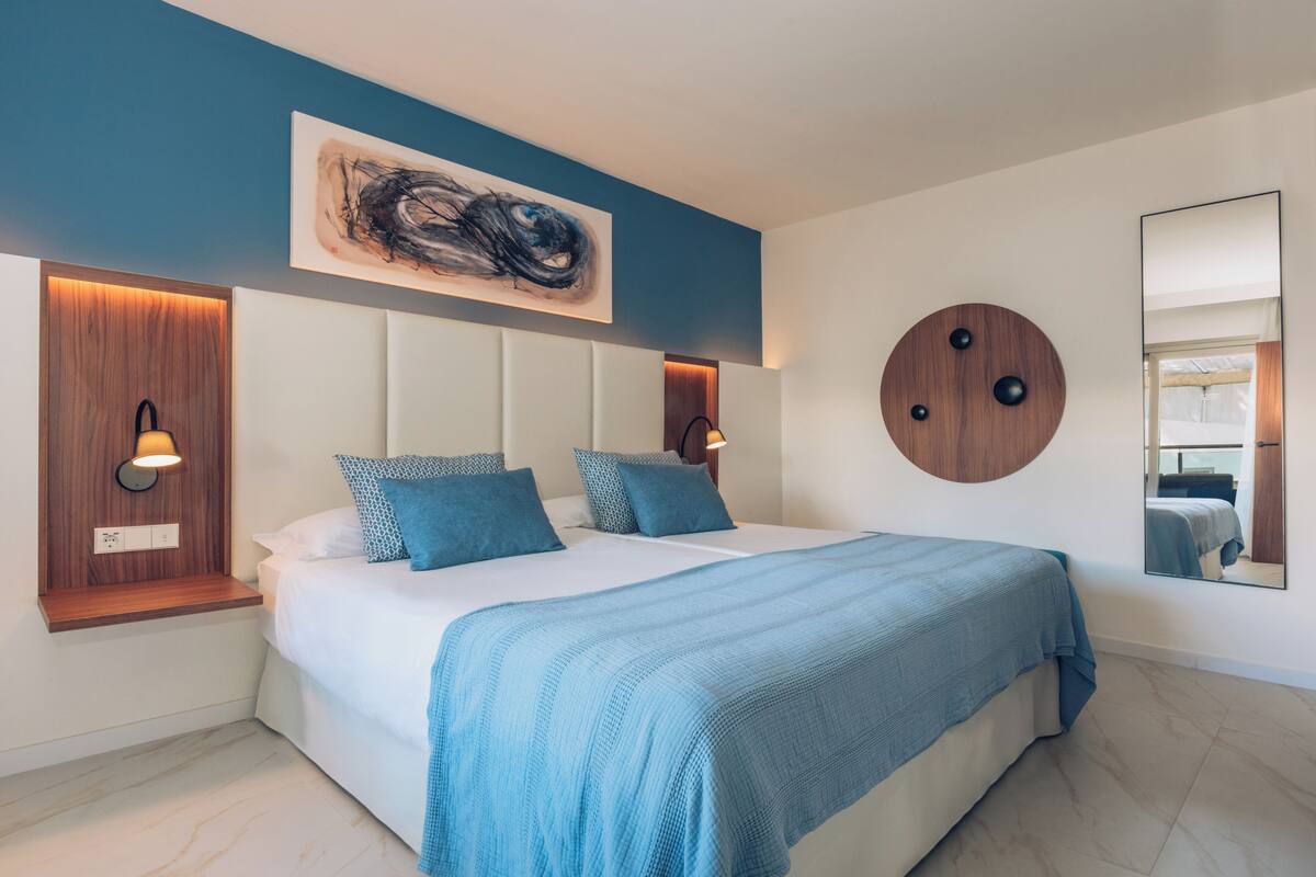 Une chambre moderne avec un grand lit, un mur d’accent bleu, un élément décoratif rond en bois et un grand tableau abstrait.