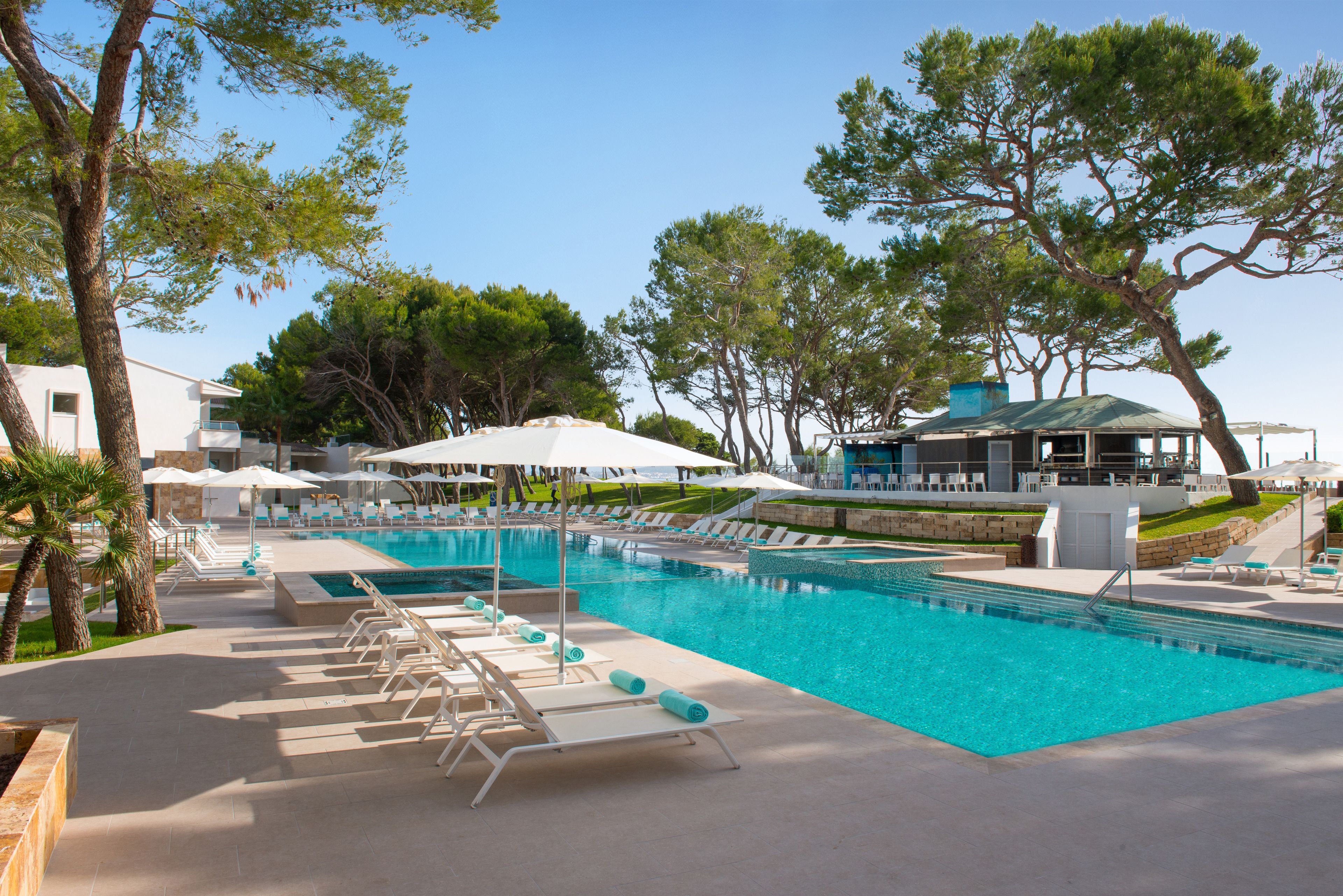 A swimming pool with lounge chairs and umbrellas, surrounded by trees and a building with a green roof.