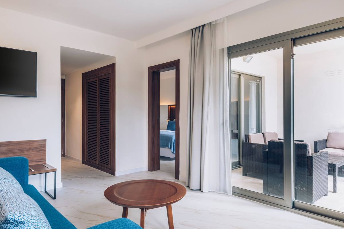 Une chambre d’hôtel moderne avec un canapé, une petite table en bois, un grand miroir et une porte-fenêtre coulissante donnant sur un balcon aménagé avec du mobilier d’extérieur.