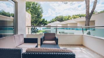 Un balcon doté de meubles en osier, d’une rambarde en verre et offrant une vue sur une piscine et des immeubles.