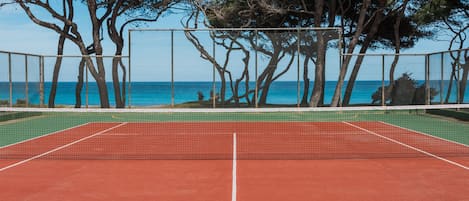 A tennis court with a net, surrounded by trees and a view of the sea.