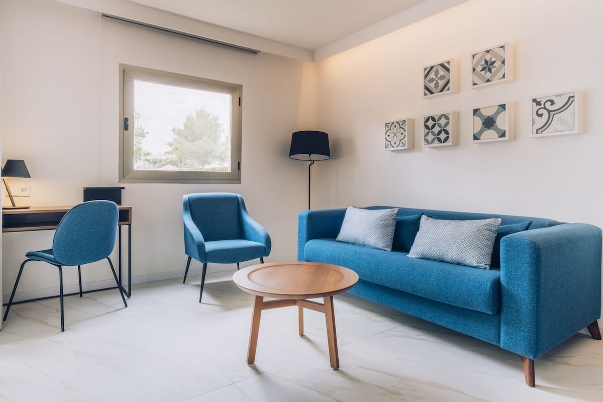 Un salon moderne avec un canapé bleu, deux fauteuils assortis, une table basse en bois, un bureau avec une lampe et une fenêtre.
