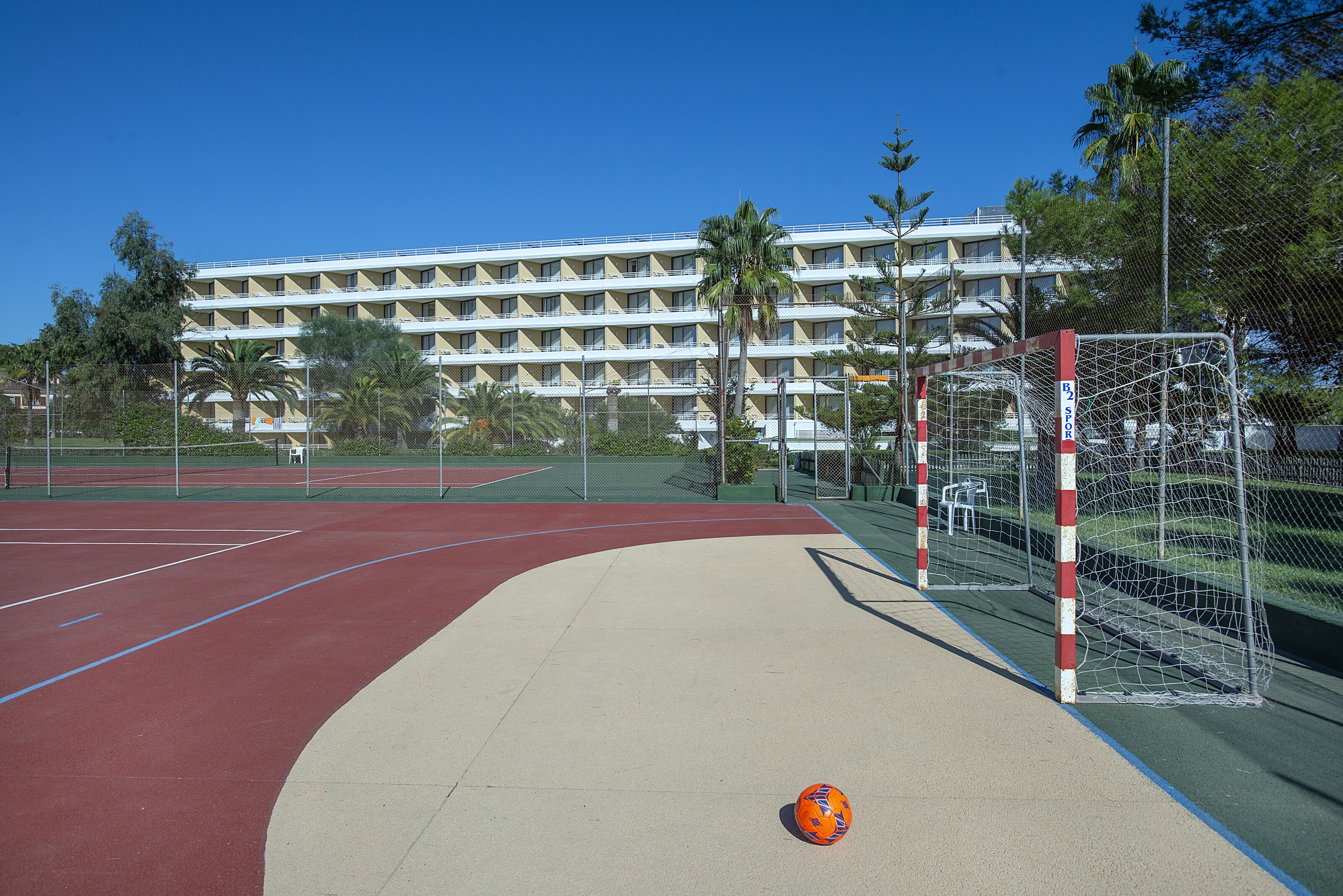 Una cancha de baloncesto al aire libre con un aro y una pelota, rodeada por una valla y palmeras, con un edificio al fondo.