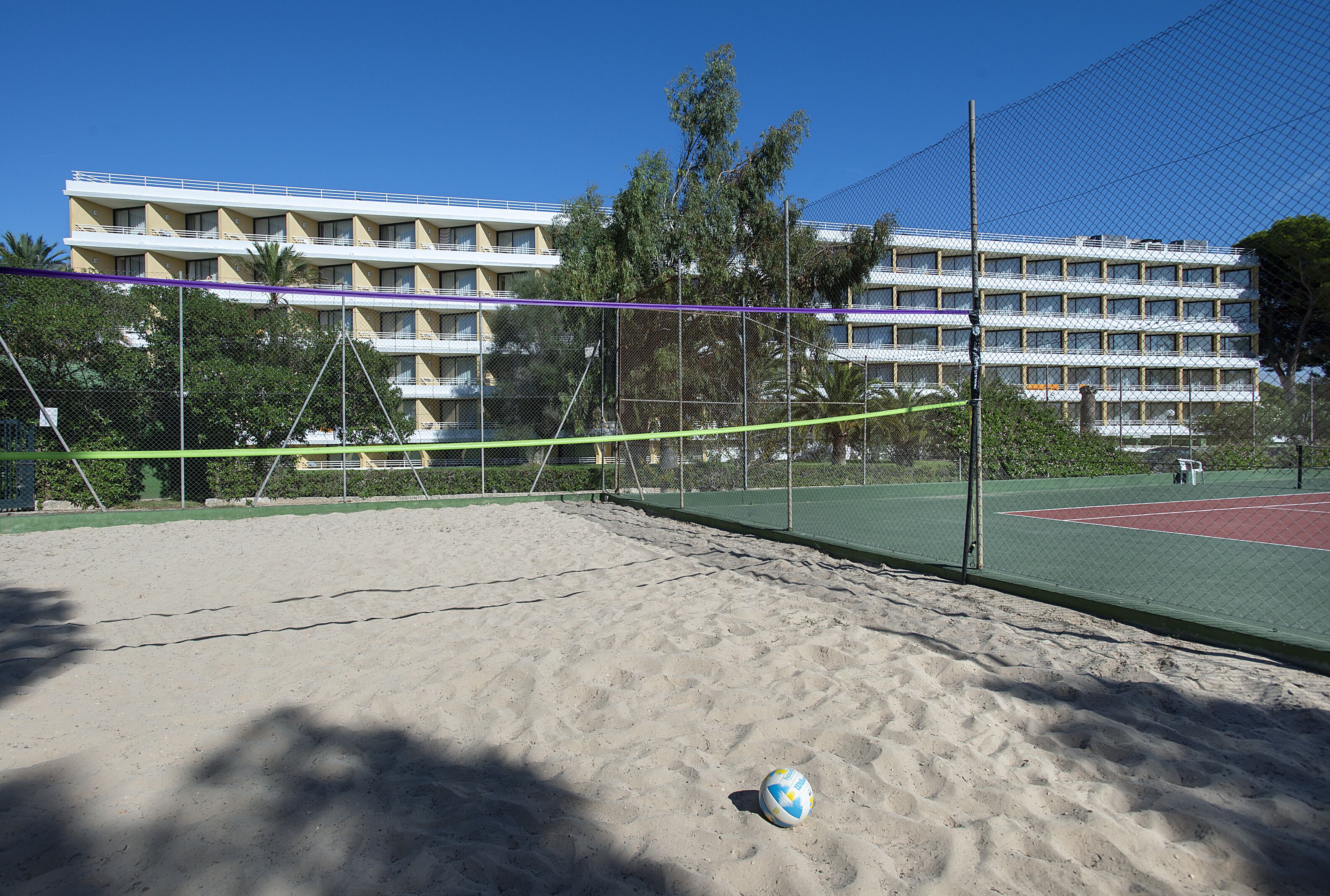 A sandy beach volleyball court with a net, surrounded by a fence, and a building with multiple balconies in the background.