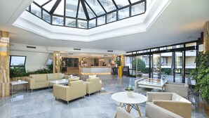 A spacious lobby with a high ceiling and skylight, featuring a reception desk, seating area with sofas and chairs, and a view of the outdoor balcony. - Hotel Exagon Park (Santa Margalida)
