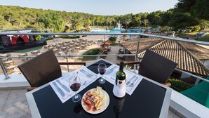 A table with a bottle of wine, glasses, and a plate of food on a balcony overlooking a pool area with lounge chairs and umbrellas. - Hotel Exagon Park (Santa Margalida)