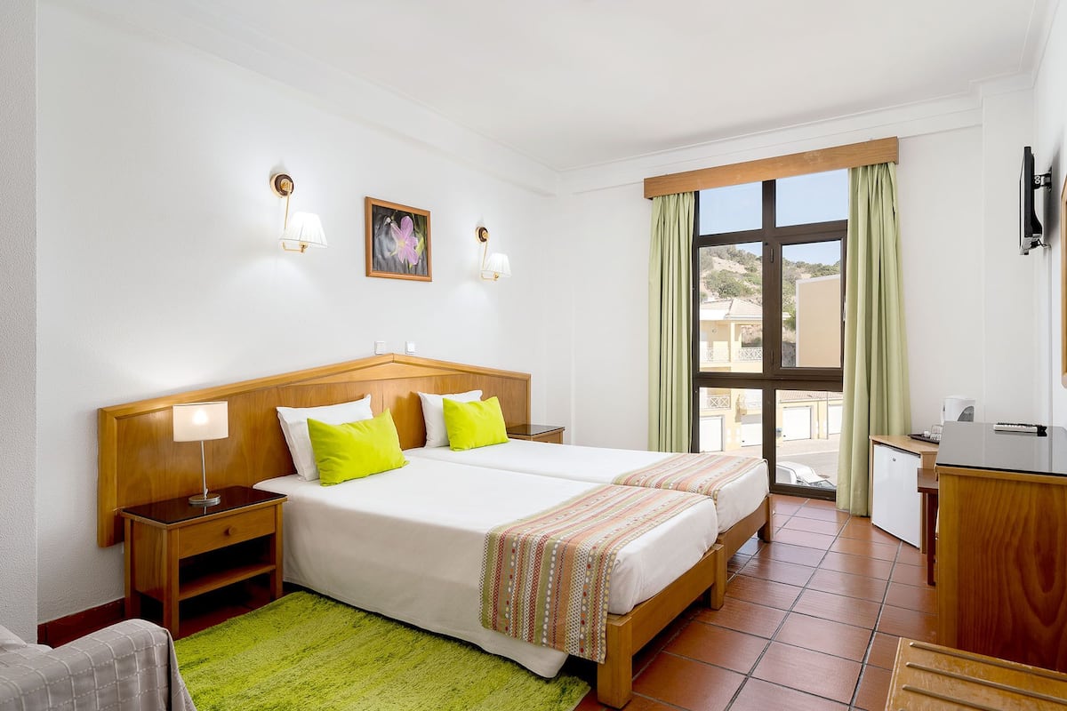 A hotel room with two beds, a desk, a chair, a green rug, and a window with curtains.