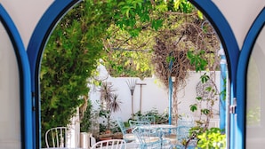A view through a blue-framed window of an outdoor seating area with white chairs and tables, surrounded by greenery and plants. - Nathalie Hotel (Rhodes)
