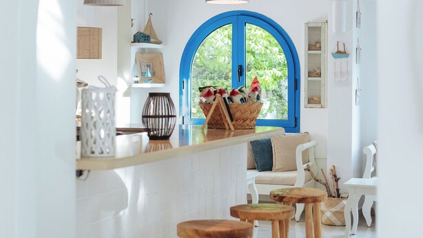 A kitchen with wooden stools, a window with a blue frame, and hanging pendant lights. - Nathalie Hotel (Rhodes)