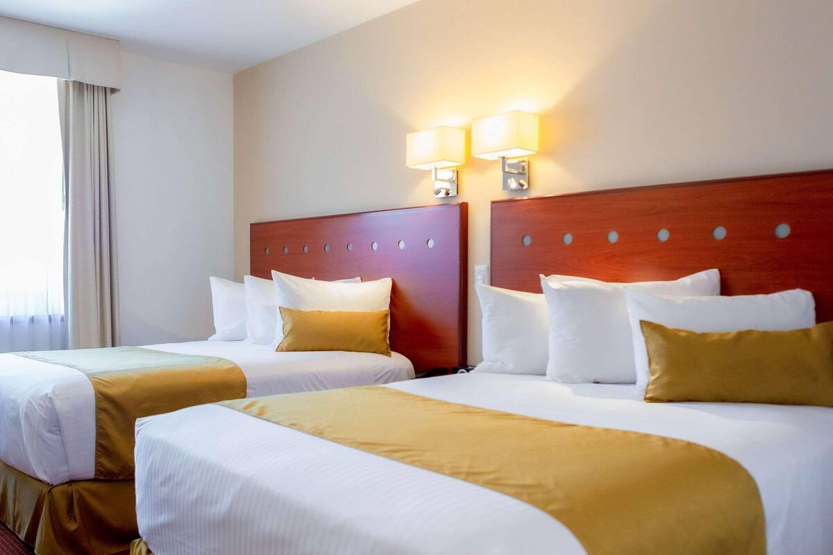 A hotel room with two beds, a wooden headboard, two wall-mounted lamps, and a window with curtains.