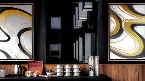 Un coin bar avec une machine à café, une pile de tasses blanches et un tableau abstrait encadré accroché au mur.