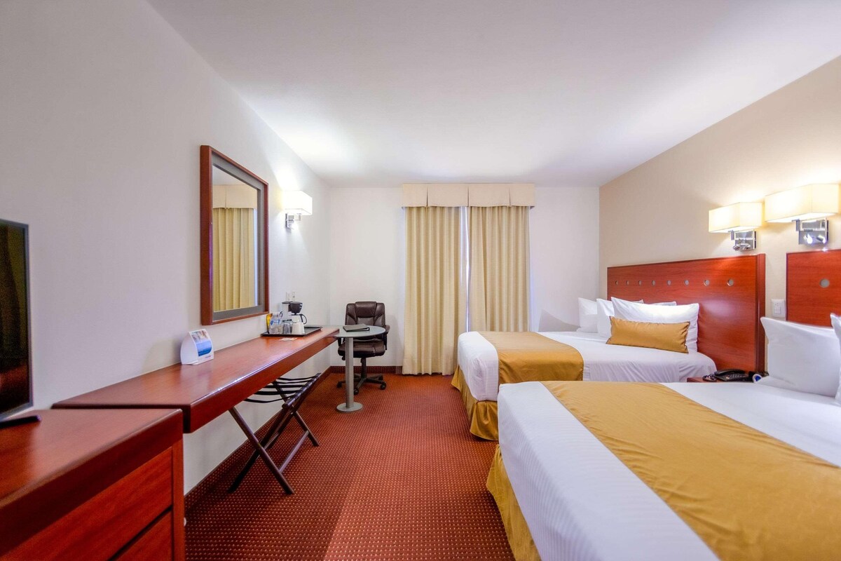 A hotel room with two beds, a desk, a chair, a mirror, and a window with curtains.