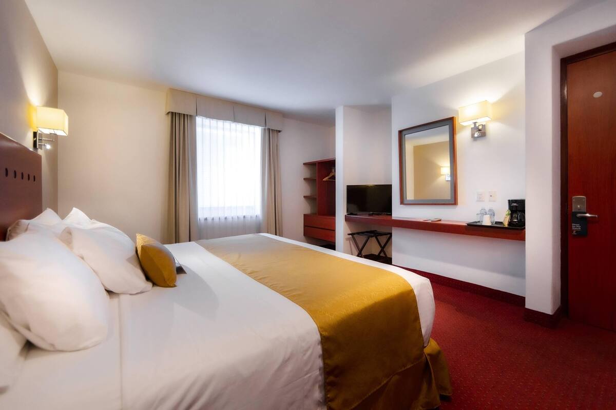 A hotel room with a large bed, a desk with a computer, a mirror, and a television.