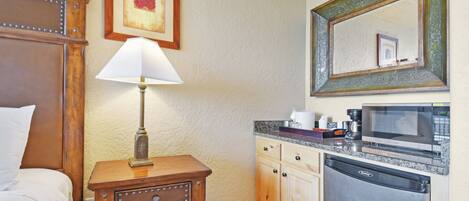 A hotel room with a bed, bedside table with a lamp, a wooden nightstand, a kitchen area with a microwave, a dishwasher, and a framed picture on the wall.