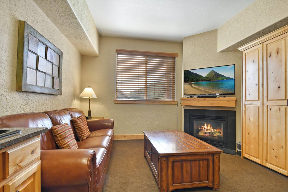 A living room with a leather sofa, a wooden coffee table, a fireplace, a television mounted above it, and a window with blinds.