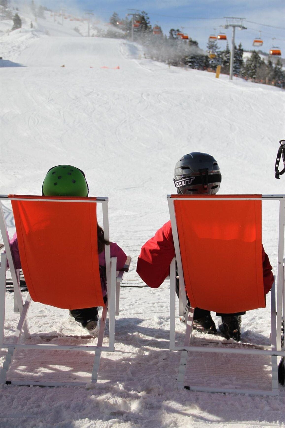 Two children in ski chairs on a snowy slope with ski lifts in the background.