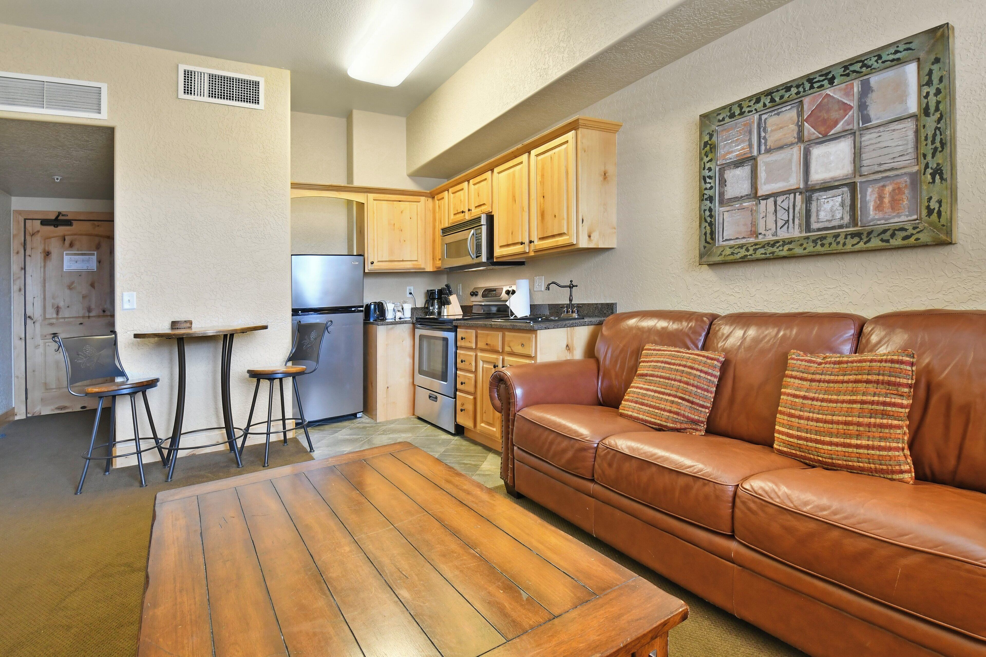 A kitchen with wooden cabinets, a microwave, and a refrigerator. There is a brown leather sofa with pillows and a wooden coffee table.